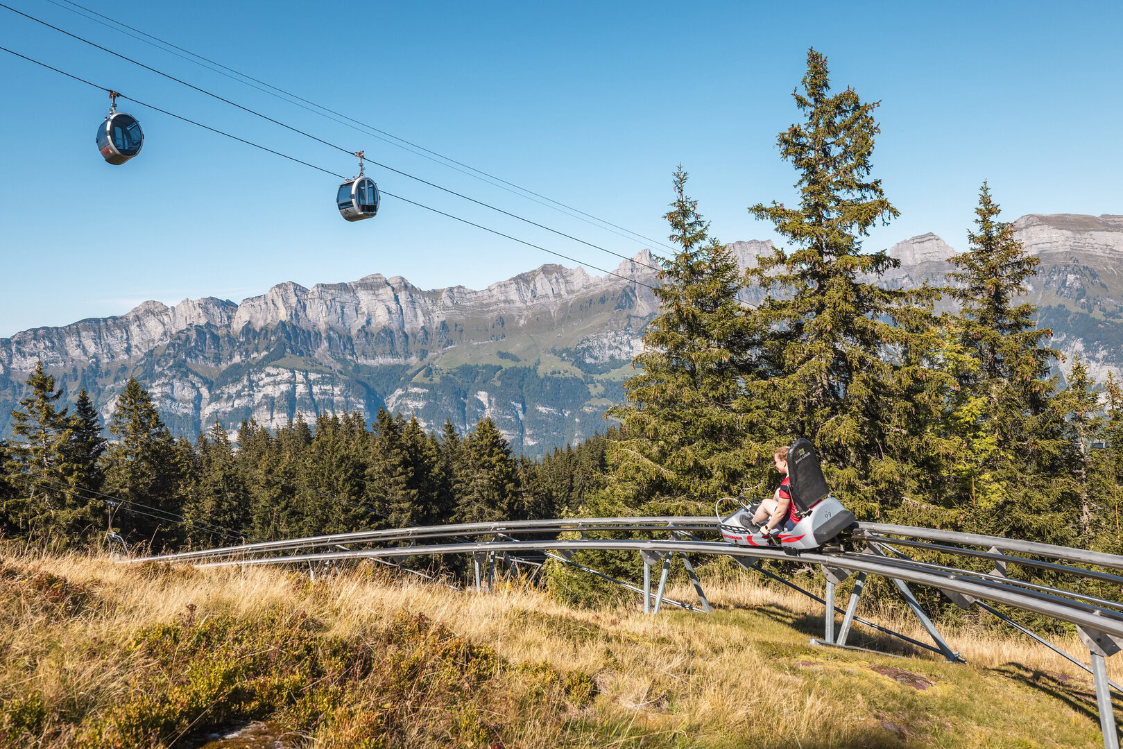 Person, die an einem sonnigen Tag mit einer Bergachterbahn fährt, über deren Köpfen Seilbahnen schweben, umgeben von immergrünen Bäumen und einer Kulisse aus schroffen Bergen.