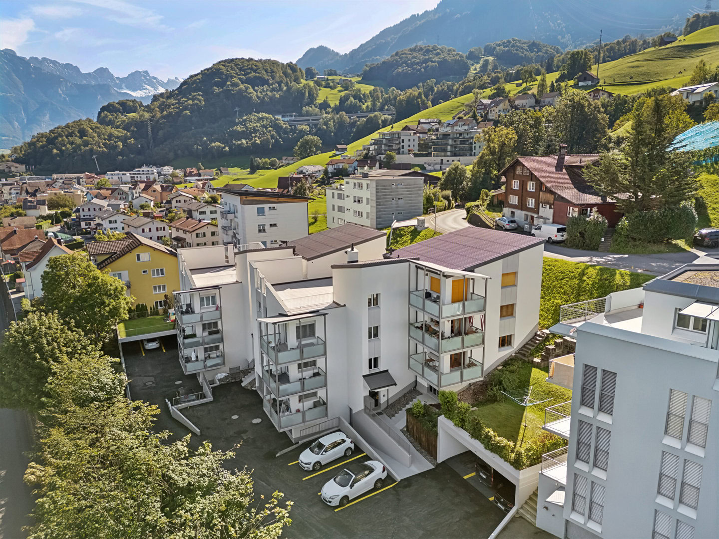 Luftaufnahme eines Wohngebiets mit modernen Wohnhäusern, umgeben von grünen Hügeln und Bergen im Hintergrund.