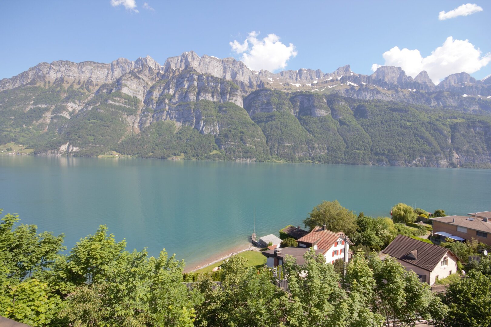 See mit klarem, blauem Wasser vor der Kulisse felsiger Berge und einer Küstenlinie mit Häusern und Bäumen im Vordergrund.