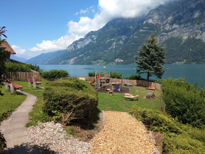 Ein Spielplatz am Seeufer mit Rutschen und Sandkasten, umgeben von üppigem Grün. Im Hintergrund sind Berge und Wolken zu sehen, ein Holzweg führt zum Spielbereich.